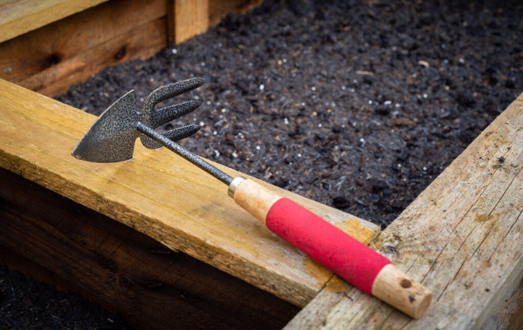 Wood and steel double sided hoe and rake garden hand tools, resting on top of a freshly planted wooden vegetable planter box full of soil in a rustic backyard residential garden.